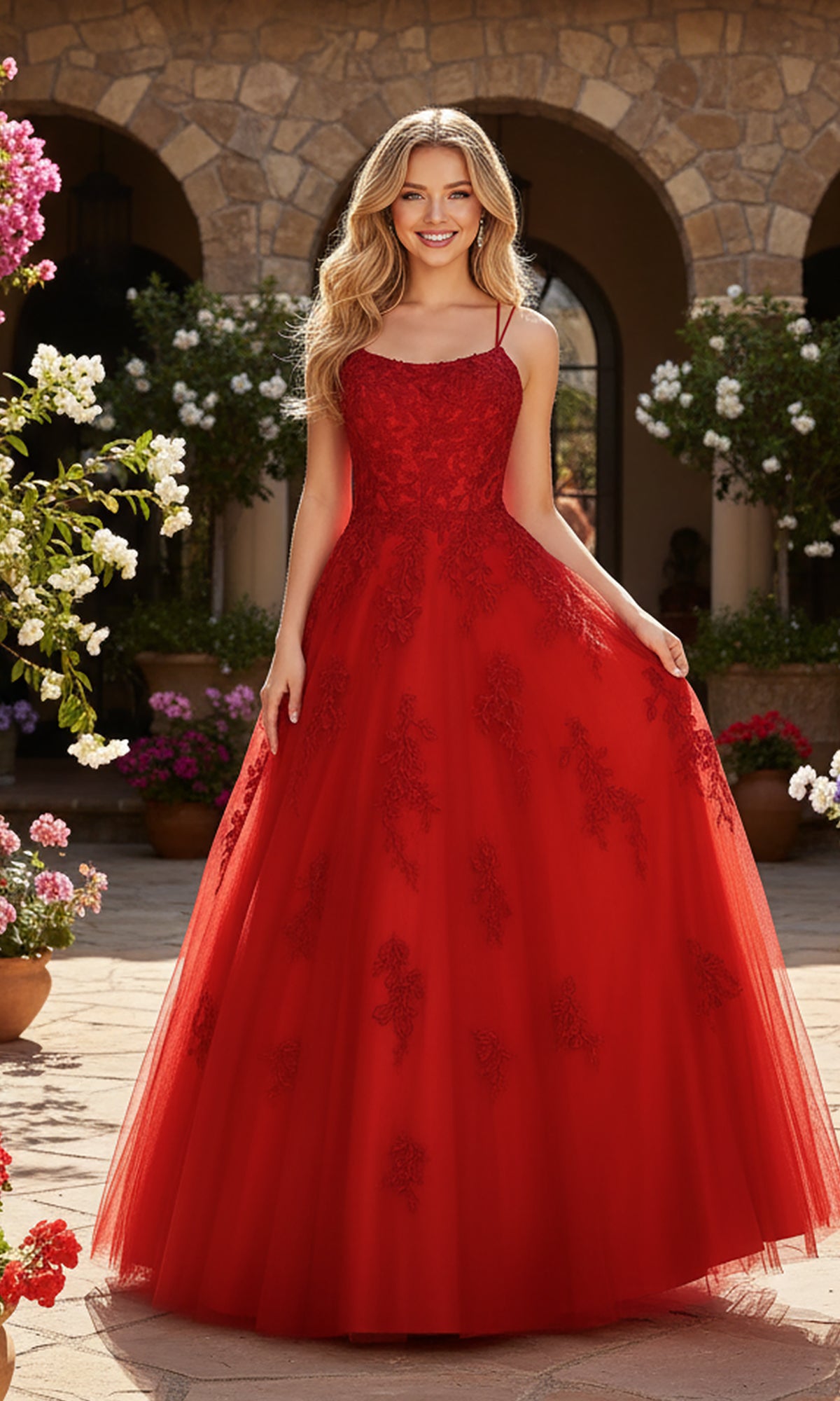 A woman stands outdoors in the PromGirl Exclusive 250228, a long red lace prom dress with an adjustable lace-up back and thin straps, surrounded by potted flowers and an arched stone building.